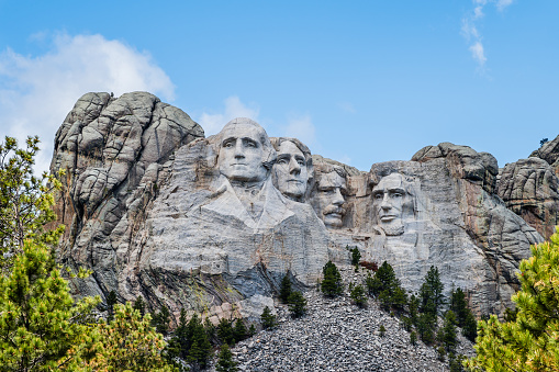 Front view of Mount Rushmore. The carved faces of the four historical figures, George Washington, Thomas Jefferson, Theodore Roosevelt and Abraham Lincoln are framed by a brilliant stormy sky and tree branches. Bright green coniferous trees in the foreground provide contrast to the granite stone.
