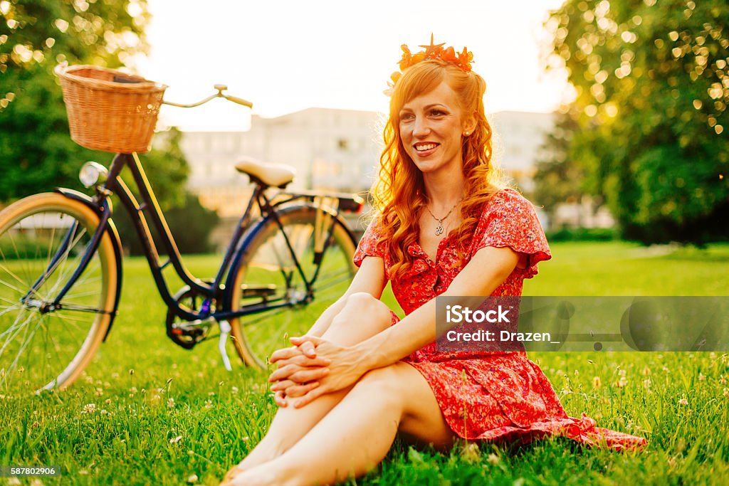Relaxed afternoon in city park Modern urban red head woman in summer dress with hair accessory. Ginger woman is riding vintage bicycle with basket on steering bar. Active Lifestyle Stock Photo