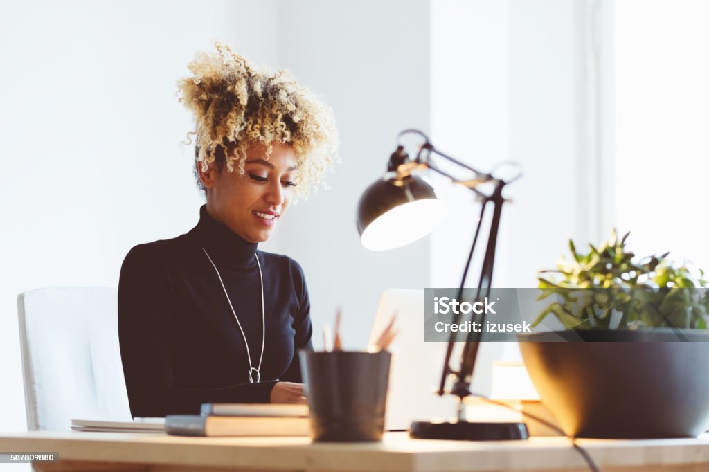 Afro american young woman using a laptop in an office Beautiful afro american elegant young woman sitting at the desk and using a laptop in an office or at home.  Working At Home Stock Photo