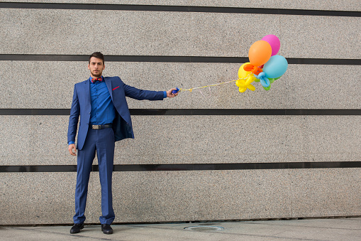 Young businessman standing outdoors and holding bunch of colorful balloons against the wall while looking at camera.
