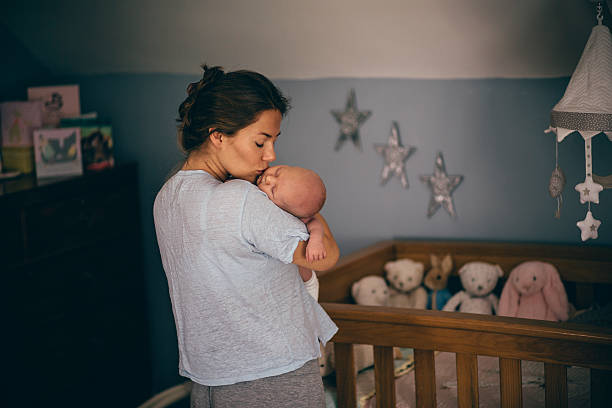 Time for bed A happy new mother kisses her newborn baby on the cheek as she gets ready to put her down in the cot to sleep. The bedroom has stars on the walls and there are soft toys in the cot. baby sleeping bedding bed stock pictures, royalty-free photos & images