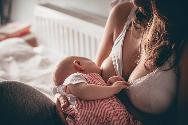 Mother Breastfeeding A mother breast feeds her newborn baby in her bedroom. She is wearing a breastfeeding bra and casual trousers while her little girl is dressed in red striped dungarees. bra stock pictures, royalty-free photos & images