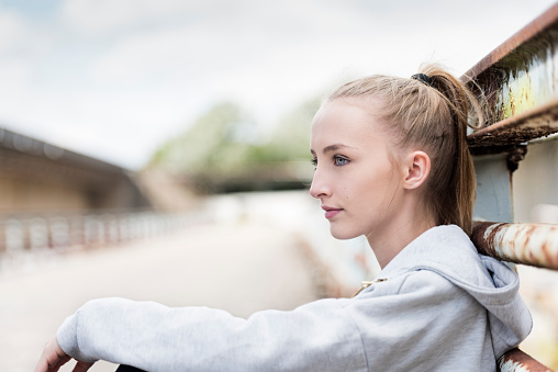 Portrait of an athletic looking young woman resting before she starts her workout. She is sitting on the floor with her back against an old rustic fence. Photographed at her eye level with her face turned away from the camera in profile with a shallow focus effect with the point of focus being on her eye. She is wearing a grey tracksuit top and she has fair coloured hair pulled back in a pony tail and a fresh clean complexion. Colour, horizontal format with lots of copy space in the background behind her.