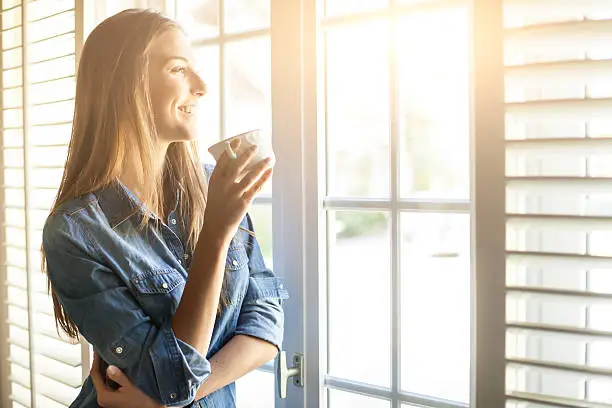 Cheerful young woman drinking coffee and looking through window. With long hair and denim shirt. Tall windows with shutters, sunbeam.