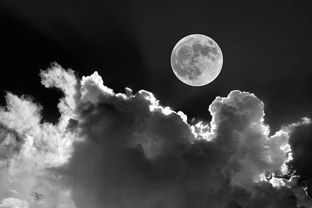 Black and white photo of a full moon over fluffy white clouds illuminated beautifully against the night sky.
