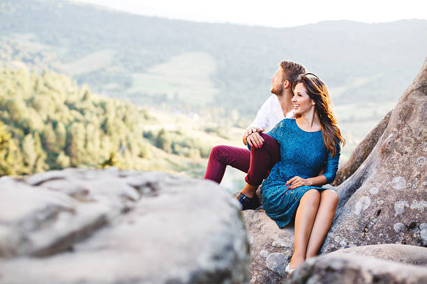 Nice couple sitting on high rock, outdoor. Girl leaning on her boyfriend's knee. Man putting his hand on girl's hand. Beloved looking afield and smiling. Woman wearing blue dress and man wearing white shirt, black shoes and claret trousers. Profile
