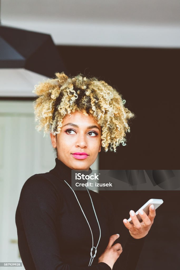 Afro american young woman holding a smart phone Beautiful afro american young woman holding a smart phone in hand indoors. Smart Phone Stock Photo
