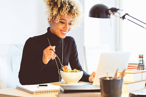 Afro american young woman eating lunch at the desk Beautiful afro american young woman sitting at the desk in an office and eating lunch, using laptop at the same time. lunch stock pictures, royalty-free photos & images