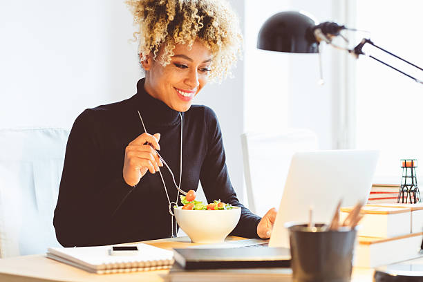 afro amerykańska młoda kobieta jedząca lunch przy biurku - wireless technology working at home one person reading zdjęcia i obrazy z banku zdjęć