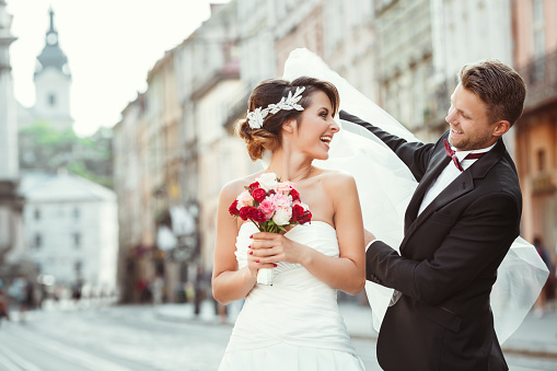 Wedding photo shooting. Bride and bridegroom walking in the city, looking at each other, holding bouquet and smiling. Groom holding veil of bride. Outdoor, cobbled street