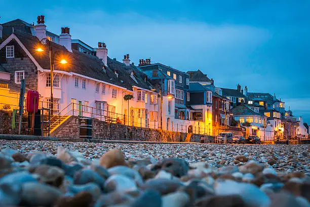 Seafront cottages warmly illuminated against the blue dusk sky overlooking the pebble beach at Lyme Regis, the historic seaside holiday resort in Dorset, UK.