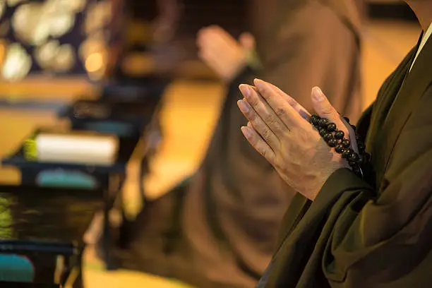 In front view, close up of monk's hands in prayer with a rosary. Visible just hands. In background blurred image of other monk kneeling in prayer. Chion-ji temple in Kyoto, Japan