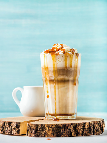 Latte macchiato with whipped cream and caramel sauce in tall glass on round wooden serving board over blue painted wall background, selective focus, vertical composition