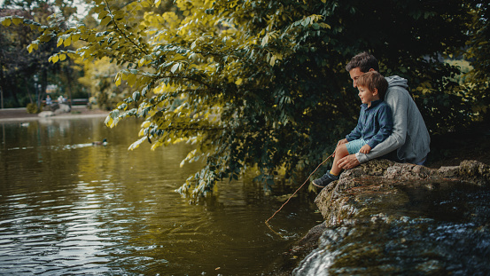Little smiling boy is learning how to catch a fish in a park pond, with a little help of his father; wide photo dimensions