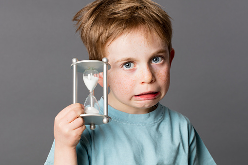 scared little child with red hair showing an hourglass, showing his worry in growing up for time concept, grey background