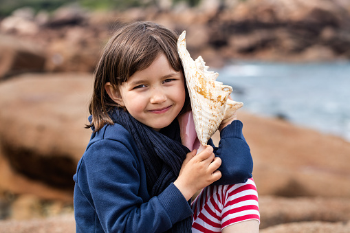 smiling beautiful young kid listening to the ocean, holding a seashell for holiday memories, relaxing for her summertime or springtime vacation on the sea