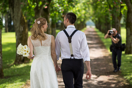 Bride holding bouquet of flowers and walking with groom on single track in park, photographer taking photo with digital camera seen in background.