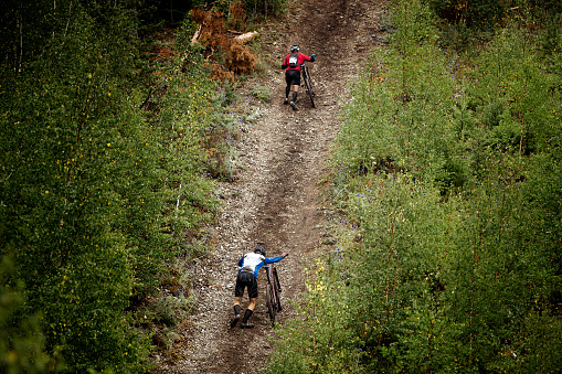 Revda, Russia - July 31, 2016: two athlete mountainbiker walking with bicycle uphill during Regional competitions on cross-country bike