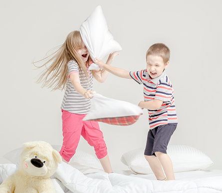 Two cute young children (boy and girl) dressed in casual clothes are playing having fun on white bed. The cheerful brother and the blonde elder sister are fighting pillows. Studio shooting on white background