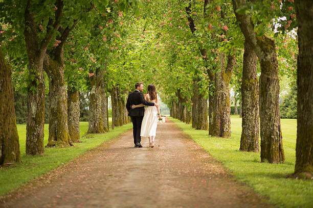 Bride holding bouquet and walking with groom on single track in park.