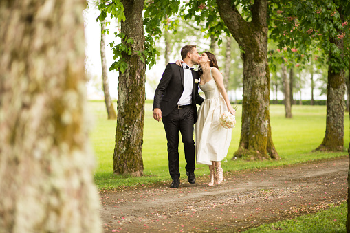 Bride holding bouquet and kissing to groom while walking on single track in park.