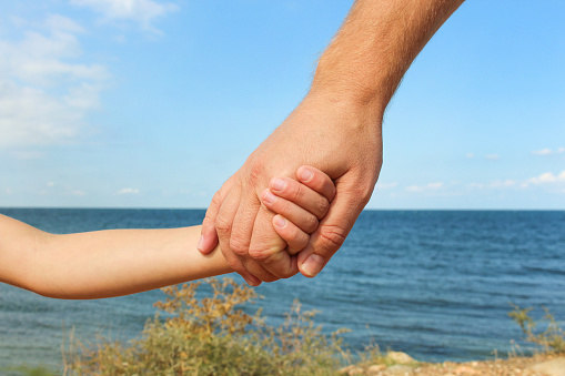 Man holds hand of child on background of sea and sky Concept of love, care, friendship, trust in family.