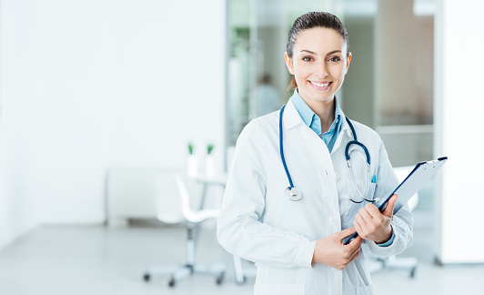 Smiling female doctor with lab coat in her office holding a clipboard with medical records, she is looking at camera