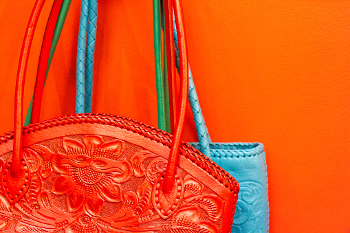 Traditional Mexican leather purses (orange-red and blue) hang against a vibrant orange-red wall.  Copy space available.