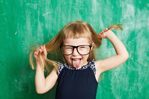 Little child with mischief looking at camera. Cheerful brat holding hair and sticking tongue out. Cute girl wearing eyeglasses. Its photo illustrating childhood. It is perfect for using it in commercial and advertising photography, reports, books, presentation