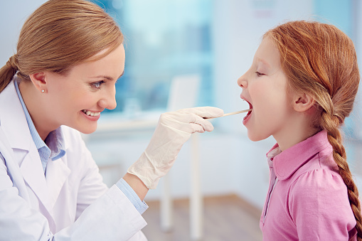 Side view of kind doctor examining throat of child. She treating tonsillitis. Redhead girl with open mouth looking at general practitioner. Its photo illustrating medical examination and healthcare and medicine. It is perfect for using it in commercial and advertising photography, reports, books, presentation