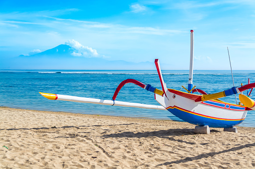 A traditional Indonesian outrigger canoe still used for day to day fishing by locals sits on the beach on a beautiful blue sky day.