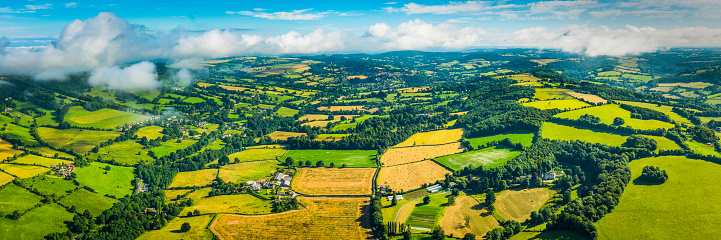 Aerial view from above the clouds over picturesque patchwork landscape of green pasture, lush meadows and golden crop fields, farmhouses and rural homes amongst the rolling hills and quiet valleys below panoramic summer skies.