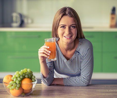 Attractive girl is holding a glass of juice, looking at camera and smiling while standing in her kitchen at home