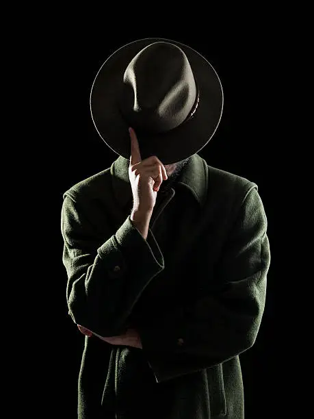 Low key portrait of man hiding his face with fedora hat in dark.He is wearing a dark green coat.The light source is behind model.He is touching his hat while obscuring his face.Isolated on black with clipping path.Shot in studio with medium format DSLR camera Hasselblad.