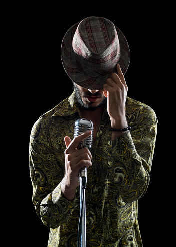 Portrait of hipster man wearing an old fashioned shirt and hat singing in dark via microphone.He is holding microphone with right hand and touching his hat with left hand.Isolated on black with clipping path.Shot in studio with medium format DSLR camera Hasselblad.