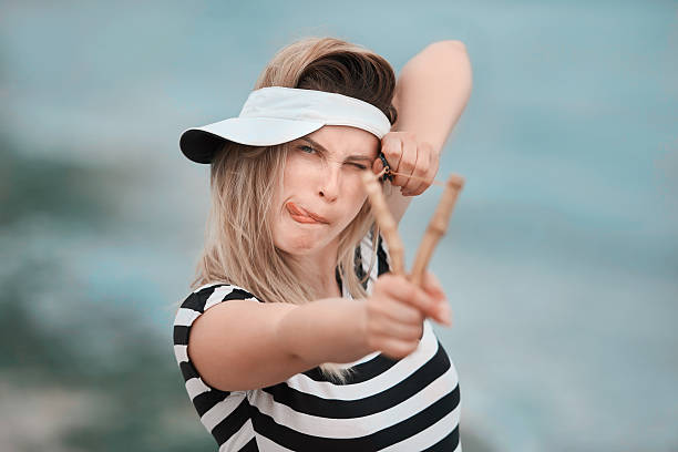funny and grimacing woman sticking out tongue while aiming to the camera with her slingshot, fun concept.
