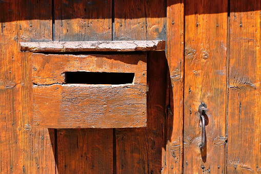 Horizontal composition brown, orange color photography of close-up in front view of old, textured and weathered domestic mailbox hanging on very old wooden door. This letterbox is homemade, all in wood material, stained with brown paint, and taken under sunlight with some shadow in summer season in a small french village outdoor in Europe. There is an old metalic handle, handful for opening the gate on right of the picture.