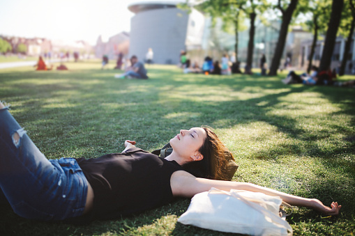 Vintage toned image of a young woman, lying on the grass, relaxing in the beautiful summertime day in an Amsterdam, Holland city park. She is wearing a casual outfit, a shirt and jeans, lying barefoot on the ground, daydreaming carefree.