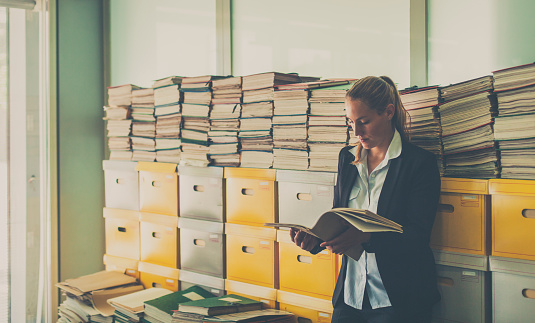 Young businesswoman working in the archive. She is looking at a file while leaning on a stack of archive boxes and documents.