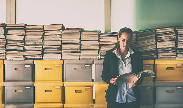 Young businesswoman working in the archive. She is looking at a file while leaning on a stack of archive boxes and documents.