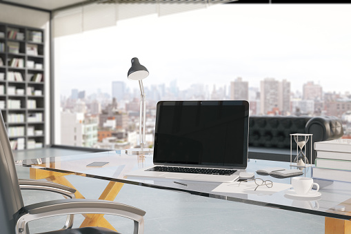 Closeup of creative office workplace with blank laptop, coffee cup, smartphone, hourglass and other items with bookshelf and window with city view in the background. Mock up, 3D Rendering