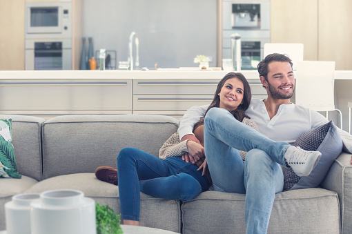Portrait of couple relaxing on the sofa. They are cozy in a luxury home. Both are casually dressed and embracing. There is a modern kitchen in the background.