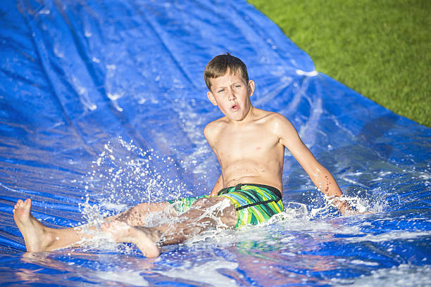 Teen boy sliding down a slip and slide outdoors Teen boy sliding down and splashing on an outdoor slip and slide. Laughing and having fun on a warm summer day greasy water stock pictures, royalty-free photos & images
