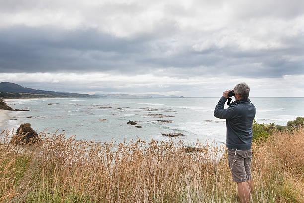 istock Gray haired man looks in binocular across ocean bay 589109642