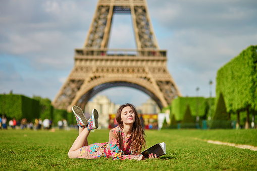 Beautiful woman in Paris lying on the grass near the Eiffel tower on a nice spring or summer day and reading a book. Student or schoolgirl doing her homework or preparing to exams