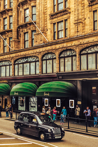 London, United Kingdom - August 4, 2016: Traditional black taxi passing by the famous Harrods department store located on Brompton Road in Knightsbridge, in the Royal Borough of Kensington and Chelsea, London.