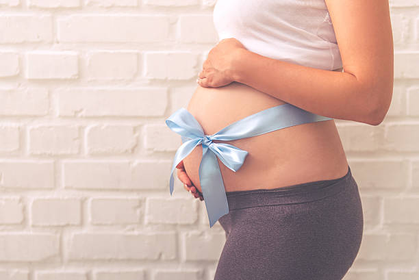Beautiful pregnant woman Side view of beautiful pregnant woman with blue ribbon on her bare tummy, on white brick wall background, cropped stomach knot stock pictures, royalty-free photos & images