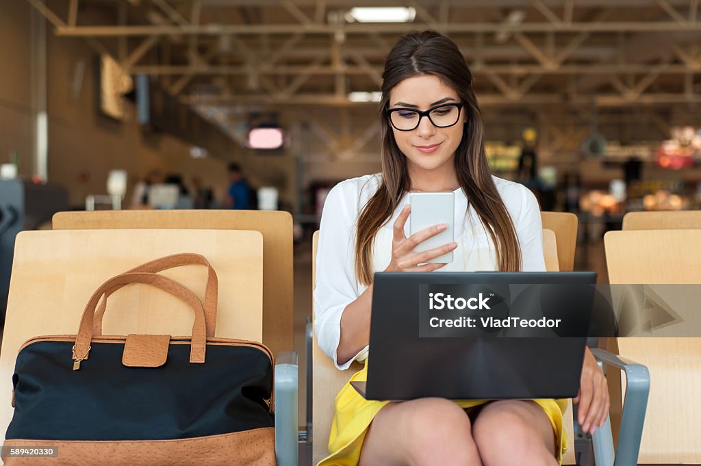 Air travel concept and modern technology Airport Young female passenger on smart phone and laptop sitting in terminal hall while waiting for her flight. Air travel concept with young casual woman sitting with hand luggage suitcase. Airport Stock Photo