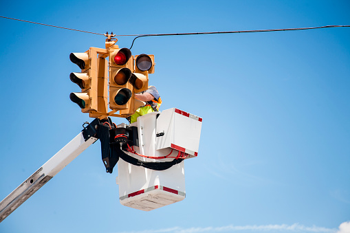 Unrecognizable utilities worker repairs a traffic signal, light using a bucket lift at a road intersection in USA.  Clear blue sky.  One man wearing safety vest.