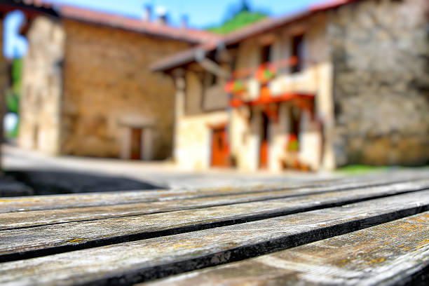 Horizontal composition color photography outdoor of selective focus on gray wooden picnic old table, plank in street of a small french village with multiple blurred stone house shapes, facade with balcony in background in summer season with a bright sunlight. Empty design background, ready with copy space with shallow depth of field.
